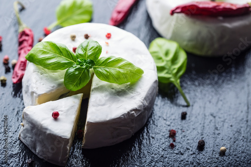 Brie cheese. Camembert cheese. Fresh Brie cheese and a slice on a granite board with basil leaves four colors peper and chili pepers. Italian and Mediterranean ingredients.