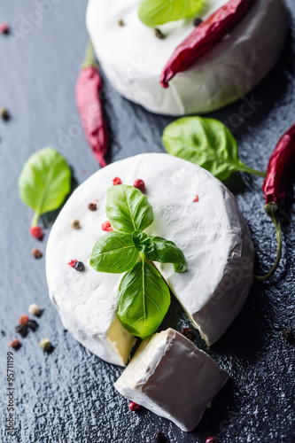 Brie cheese. Camembert cheese. Fresh Brie cheese and a slice on a granite board with basil leaves four colors peper and chili pepers. Italian and Mediterranean ingredients.