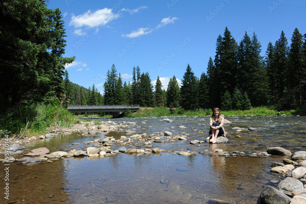 Barefoot woman enjoys the peaceful Gallatin River in the Gallatin Valley in Montana.  She is barefoot and wearing a tan jacket.