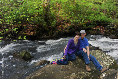 Older couple pause to rest during their hike around Lake Powhatan in North Carolina. They are both sitting on a rock besides a stream.