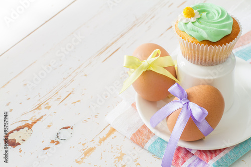Eggs decorated with ribbon and cupcakes on white wood background