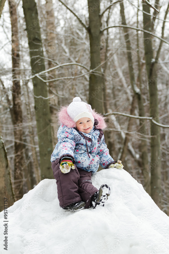 Cheerful little girl wearing warm clothes posing on snowy hill in winter forest