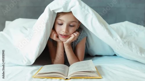 Girl child reading book under the blanket and smiling at camera