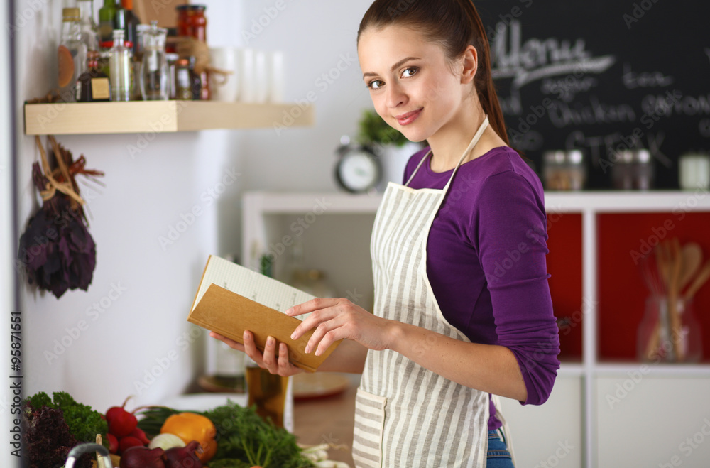 A young woman standing in the kitchen reading a cookbook.