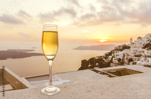 a glass of white wine on balcony with Caldera city view at sunset, yellow sunlight and bright sky before getting dark (selective focus on a glass of wine)