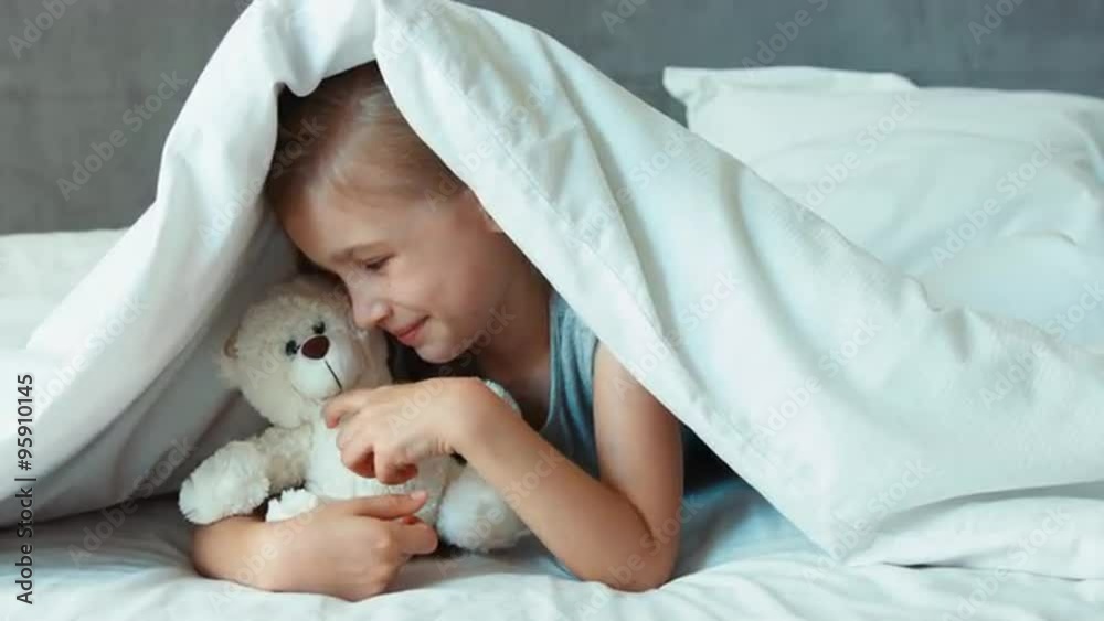 Girl hugging teddy bear under the blanket and smiling at camera Girl hugging teddy bear under the blanket and smiling at camera