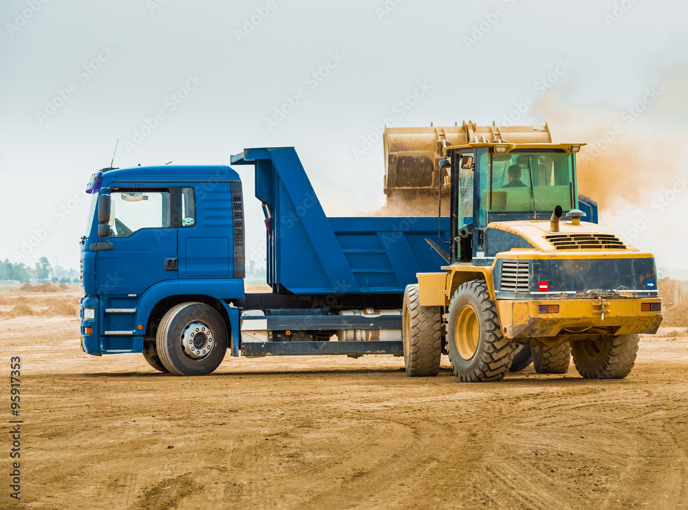 Huge backhoe loader filling the excavated soil inside blue dump truck Huge backhoe loader filling the excavated soil inside blue dump truck