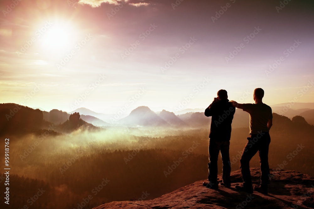 Two friends. Hiker thinking and photo enthusiast takes photos  stay on cliff. Dreamy fogy landscape, blue misty sunrise in a beautiful valley below