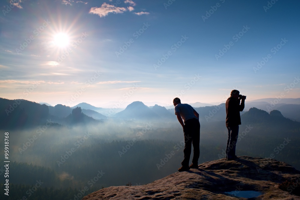 Two friends. Hiker thinking and photo enthusiast takes photos  stay on cliff. Dreamy fogy landscape, blue misty sunrise in a beautiful valley below