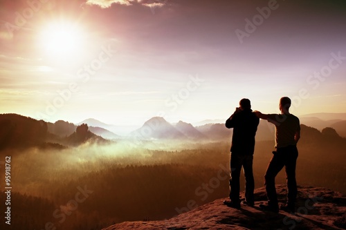Two friends. Hiker thinking and photo enthusiast takes photos  stay on cliff. Dreamy fogy landscape  blue misty sunrise in a beautiful valley below