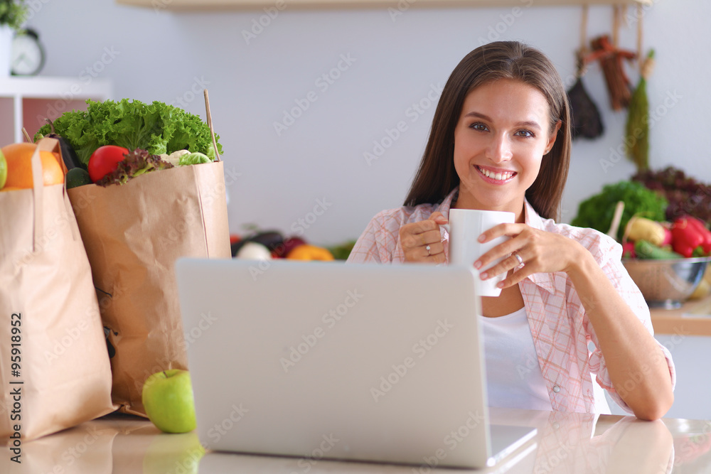 Smiling young woman with coffee cup and laptop in the kitchen at home Smiling young woman with coffee cup and laptop in the kitchen at home