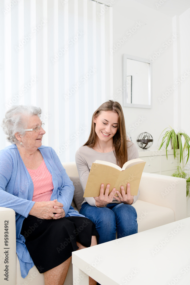 cheerful young woman reading book for elderly senior woman at home cheerful young woman reading book for elderly senior woman at home
