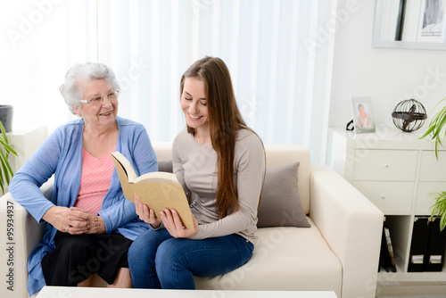 cheerful young woman reading book for elderly senior woman at home