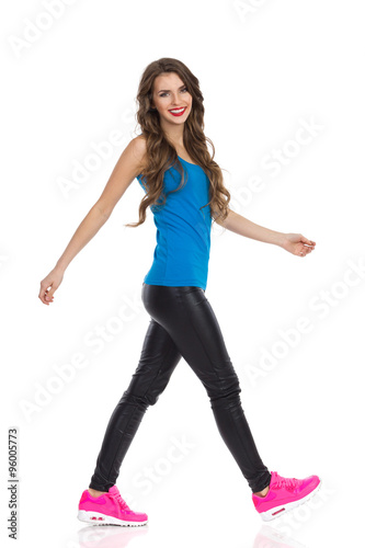Smiling young woman walking in blue shirt, black leather trousers, pink sneakers and looking at camera. Side view. Full length studio shot isolated on white.