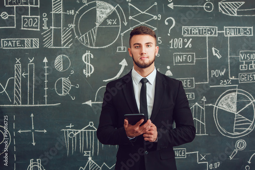 A bearded man in a suit standing with tablet near whiteboard with graphs