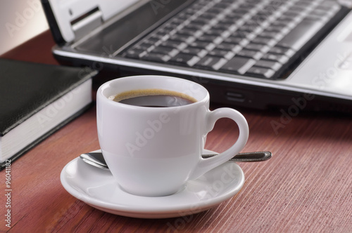 Desktop with coffee cup, opened laptop computer, diary and pen on background, no people, focused on coffee
