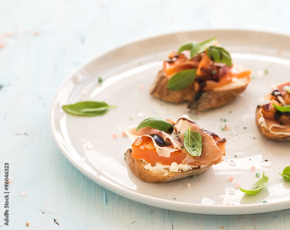Bruschettas with Prosciutto, roasted melon, soft cheese and basil on white ceramic plate over light blue background Bruschettas with Prosciutto, roasted melon, soft cheese and basil on white ceramic plate over light blue background