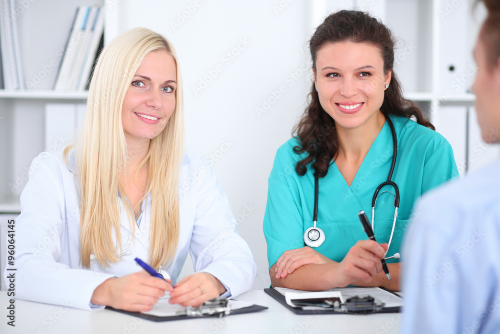 Two confident friendly female doctors sitting at the table and listen to the patient's history . Medical and health care concept Two confident friendly female doctors sitting at the table and listen to the patient's history . Medical and health care concept