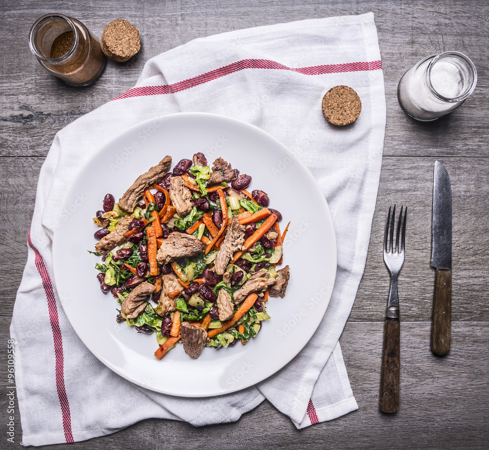 sauteed beef with grilled vegetables on white plate with knife and fork on a napkin with spices on wooden rustic background top view