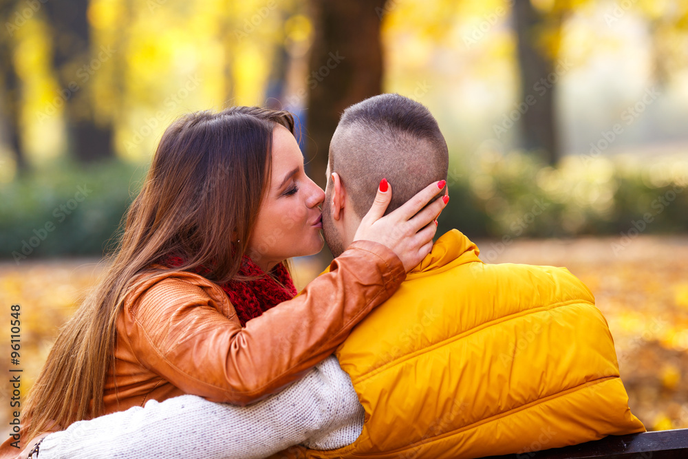 Fototapeta premium Young couple sitting on a park bench on a sunny autumn day and expressing tenderness