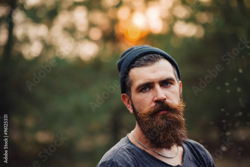 Brutal brunette bearded man in warm hat with a hatchet in the woods on a background of trees