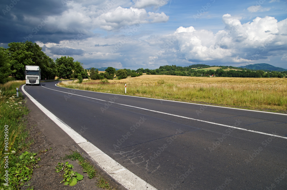 The asphalt road in rural countryside with dramatic clouds in the sky. White truck drive out from the bend between trees in the distance. Cornfield and wooded mountains in the background.