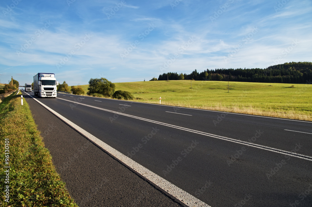 Asphalt road rising to the horizon in rural landscape. White trucks arriving from afar. Meadow and forest in the background. Sunny day with blue skies and white clouds.