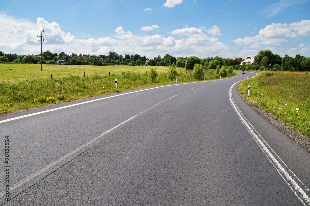 Empty asphalt road with a double bend between flower meadows in the countryside. Red car on the road in the distance. Village among the green leafy trees background. Sunny day with blue skies. Empty asphalt road with a double bend between flower meadows in the countryside. Red car on the road in the distance. Village among the green leafy trees background. Sunny day with blue skies.