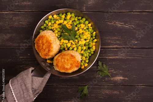 Chicken cutlets with vegetable garniture in metal skillet over wooden background, top view