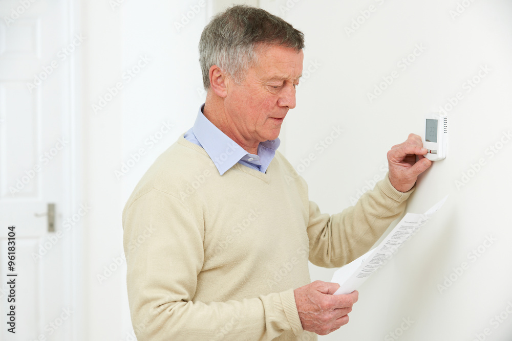 Worried Senior Man With Bill Turning Down Heating Thermostat Worried Senior Man With Bill Turning Down Heating Thermostat