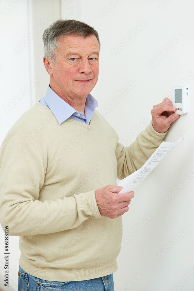 Worried Senior Man With Bill Turning Down Heating Thermostat Worried Senior Man With Bill Turning Down Heating Thermostat