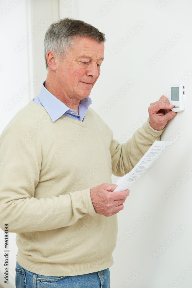 Worried Senior Man Turning Down Central Heating Thermostat Worried Senior Man Turning Down Central Heating Thermostat