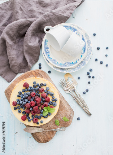 Cheesecake with fresh raspberries and blueberries on a wooden serving board, plates, cups, kitchen napkin, silverware over blue background