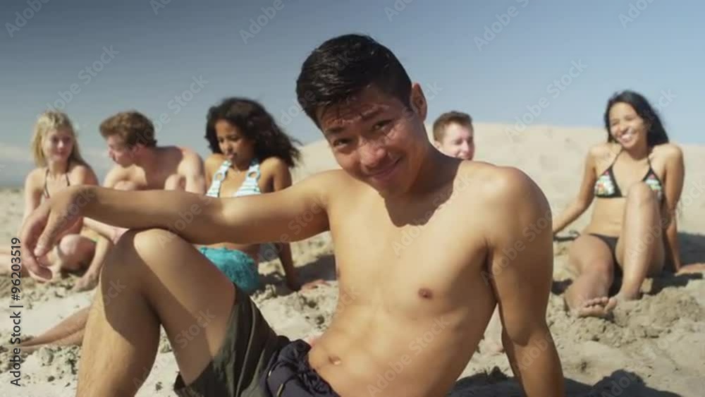 Portrait of young asian man sitting in sand with friends on the beach smiling Portrait of young asian man sitting in sand with friends on the beach smiling