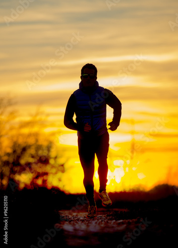Silhouette front view of young sport man running outdoors in off road trail track with Autumn sun at orange sky sunset