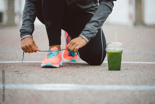 Fitness workout and healthy nutrition concept. Detox smoothie drink and running footwear close up. Female athlete tying sport shoes laces before training outdoor.