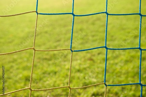Detail of yellow blue crossed soccer nets, soccer football in goal net with poor grass on playground in background.