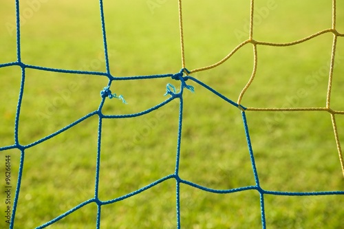 Detail of yellow blue crossed soccer nets, soccer football in goal net with poor grass on playground in background.