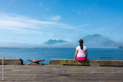 Young woman sitting on edge of dock in Tofino, British Columbia, looking out to the ocean with islands & fog surrounding them