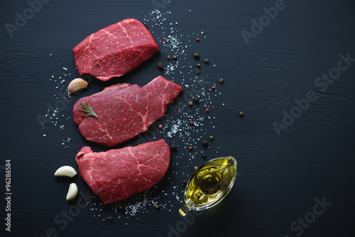 Raw marbled meat steaks with seasonings, black wooden surface photo