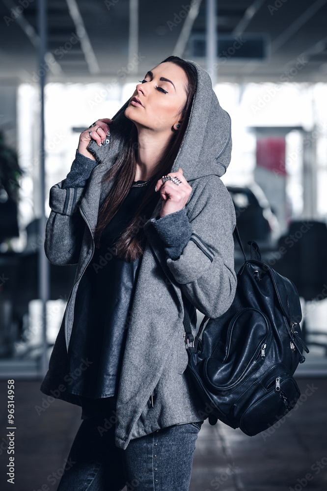 young woman with long hair, with a leather backpack, a warm hat, beautiful makeup, leather pants, a gray sweatshirt and a leather shirt, high heels walking one in the production hall and the street