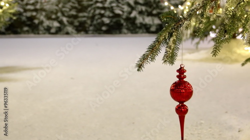 Christmas red glass finial bauble ornament swinging on spruce tree branch against white snow and distant snowy bushes background, lot of copy space.