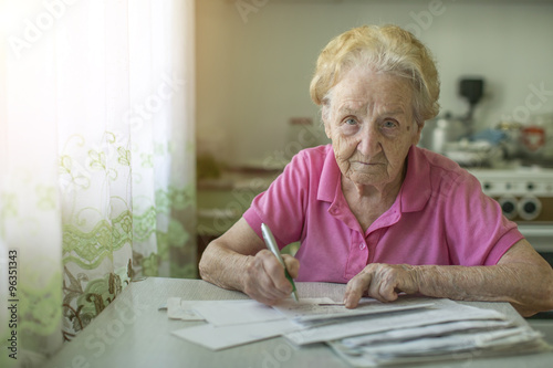 An elderly woman fills out a receipt for payment of utilities, sitting in the kitchen in house.