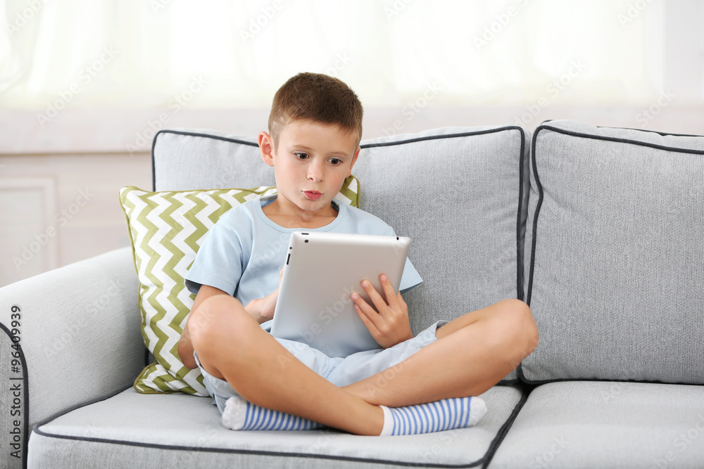 Little boy with digital tablet sitting on sofa, on home interior background Little boy with digital tablet sitting on sofa, on home interior background