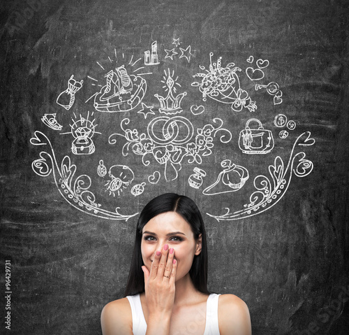 A portrait of a happy brunette lady in a white tank top who is covering her mouth by the hand and dreaming about shopping. Shopping icons are drawn on the black chalkboard.