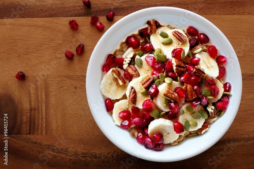 Bowl of healthy breakfast oatmeal with pomegranate, bananas, seeds and nuts, overhead view on wood