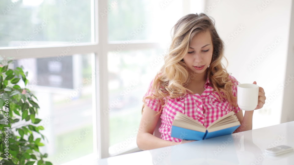 Attractive young woman sitting at table reading book and drinking from cup Attractive young woman sitting at table reading book and drinking from cup