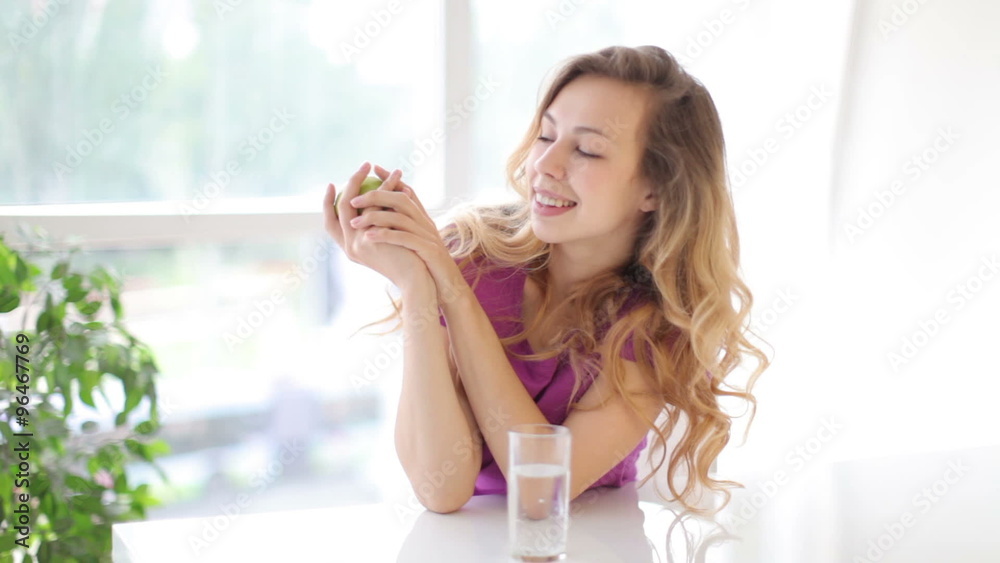 Attractive young woman sitting at table with apple in her hand and glass of water in front of her and smiling at camera Attractive young woman sitting at table with apple in her hand and glass of water in front of her and smiling at camera