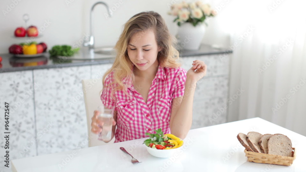 Pretty young woman sitting at table in kitchen and eating vegetable salad with smile Pretty young woman sitting at table in kitchen and eating vegetable salad with smile