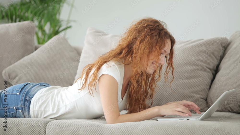 Cute young woman lying on sofa using laptop and smiling at camera Cute young woman lying on sofa using laptop and smiling at camera